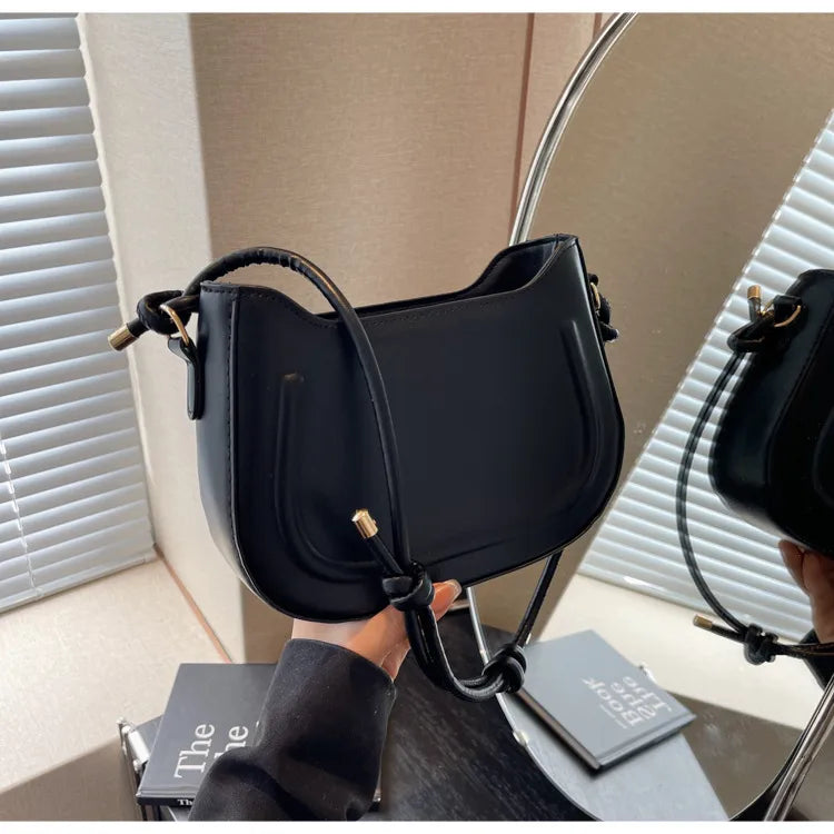 Curved shoulder bag with outlined front panel and knotted strap — black held in front of mirror | BTendance