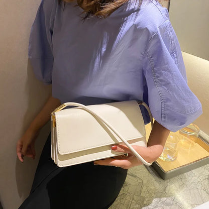 Slim rectangular flap shoulder bag with gold-tone strap accents — white held by model in purple shirt | BTendance