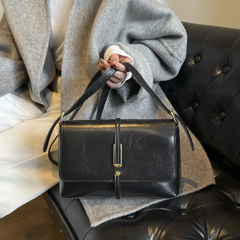 Rectangular flap shoulder bag with slim metal front detail — black held beside grey sweater | BTendance