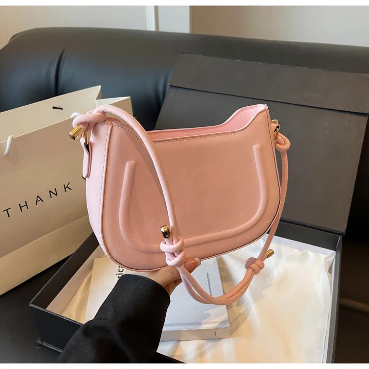 Curved shoulder bag with outlined front panel and knotted strap — pink held beside sofa | BTendance