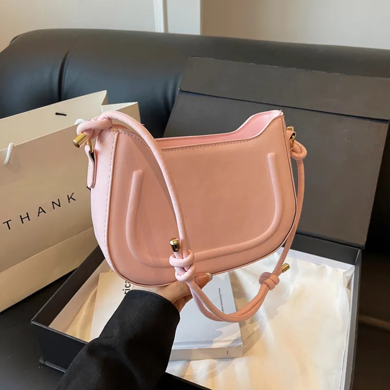 Curved shoulder bag with outlined front panel and knotted strap — pink held beside sofa | BTendance