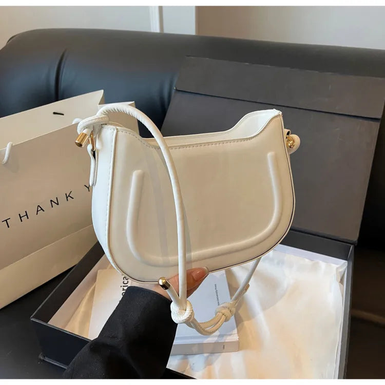 Curved shoulder bag with outlined front panel and knotted strap — cream held beside sofa | BTendance