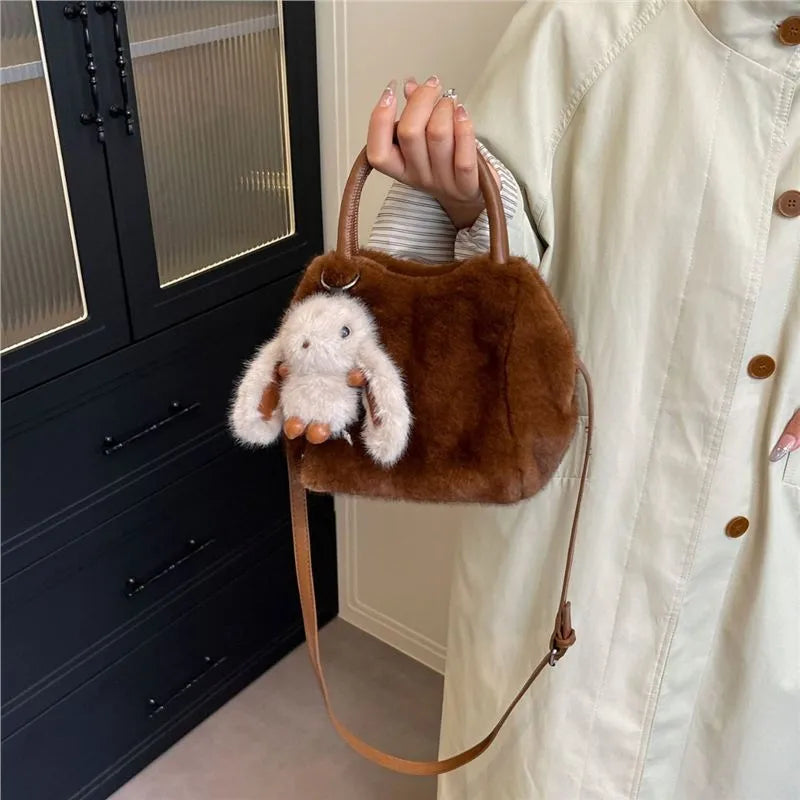 Plush top-handle handbag with rabbit charm — brown held beside cream coat | BTendance